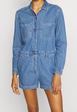 Frau trägt einen blauen Denim-Overall mit langen Ärmeln, Kragen, Fronttaschen, Gürtelschlaufen und Shorts vor einfarbigem Hintergrund.