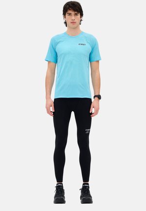 Mann steht frontal, trägt ein hellblaues kurzärmeliges Hemd, schwarze Leggings, schwarze Laufschuhe und eine schwarze Armbanduhr.