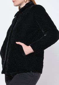 Černá sametová bunda s embosovaným květinovým vzorem, s límcem na knoflík a bočními kapsami. Vyznačuje se strukturovaným povrchem a přiléhavým střihem.