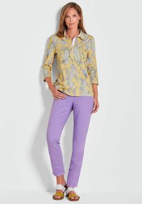 Geel overhemd met lange mouwen en bloemmotief, voorzien van een witte kraag, gecombineerd met lila broek en bruine sandalen met witte en gele accenten.