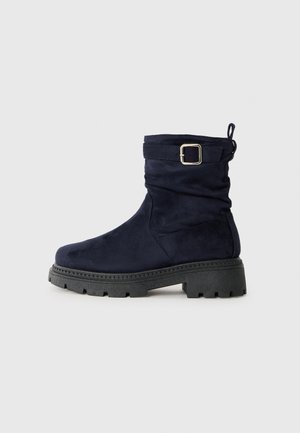 Bottes chevilles en daim bleu marine avec un design lâche, semelle épaisse noire et accent de boucle en argent sur la sangle ajustable.