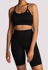 Žena v černé žebrované sportovní podprsence a vysoce pasových cyklistických šortkách, stojící na pozadí s jednolitým světlým pozadím.