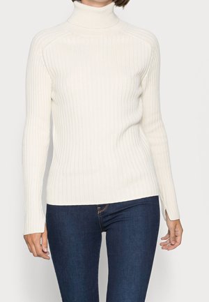 Žena má na sobě bílý žebrovaný rolák v kombinaci s tmavě modrými přiléhavými džínami na světlém pozadí.