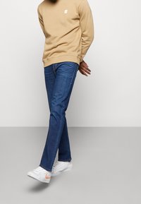 Beige sweatshirt med liten logotyp, blå jeans och vita sneakers med orangea detaljer. Avslappnad outfit med lös passform.