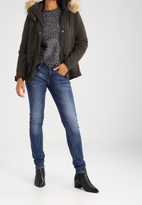 Parka verde oliva con capucha forrada de piel, combinada con un suéter de punto gris y jeans skinny azules, complementada por botas negras de tobillo.