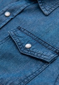 Chemise en denim avec tissu bleu, dotée d'un col pointu, de coutures contrastées et d'une poche poitrine fermée par un bouton-pression argenté.