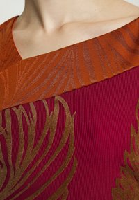 Gros plan d’un col avec un col orange rouille présentant un motif rayé structuré et un tissu côtelé bordeaux avec des motifs de feuilles marron.
