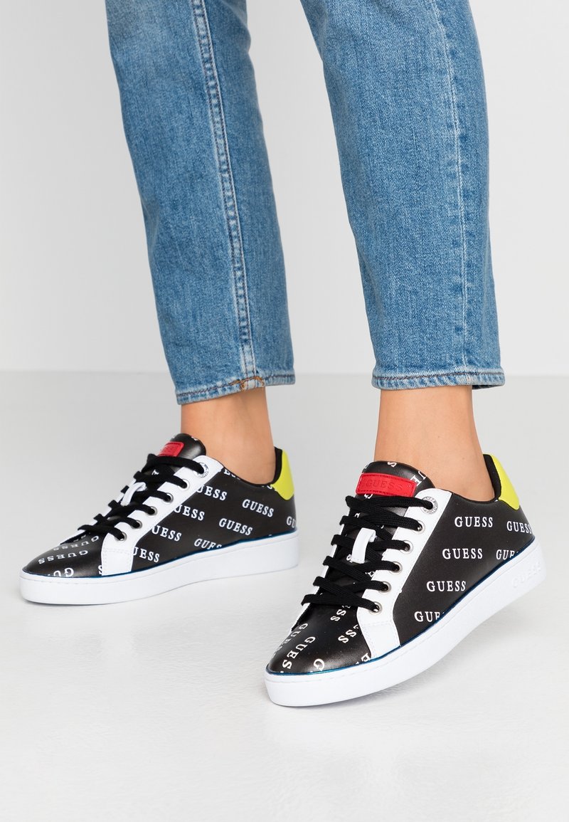Baskets noir et blanc avec une marque "GUESS", présentant un accent jaune au niveau du talon, des lacets noirs et une semelle en caoutchouc blanche.