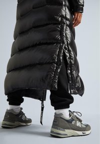 Giacca a vento nera con finitura lucida e cuciture orizzontali, abbinata a sneakers grigie New Balance con accenti riflettenti e lacci.