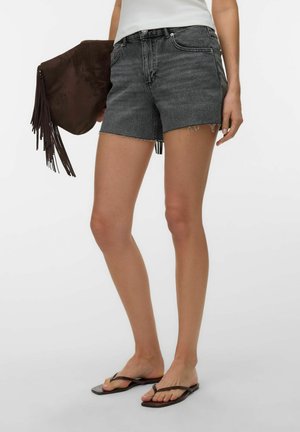 Femme portant un short en jean noir, un haut blanc, un sac à franges marron à la main et des sandales plates noires, debout devant un fond uni.