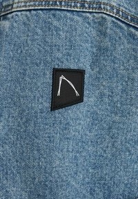 Chaqueta de mezclilla con un acabado azul texturizado que presenta un parche de tela negra con un diseño geométrico blanco.