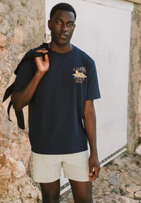 Camiseta de algodón azul marino con gráfico de "CORAL COAST", combinada con pantalones cortos beige claros y una bolsa de mano a rayas en blanco y negro. Fondo de pared texturizada.