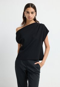 Donna che indossa un top nero a spalla unica e pantaloni neri su misura, in piedi con una mano nella tasca contro uno sfondo bianco uniforme.