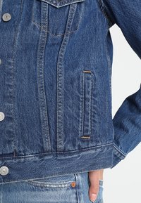 Close-up of blue denim jacket with metal buttons worn over light blue jeans, hand tucked inside front pocket.