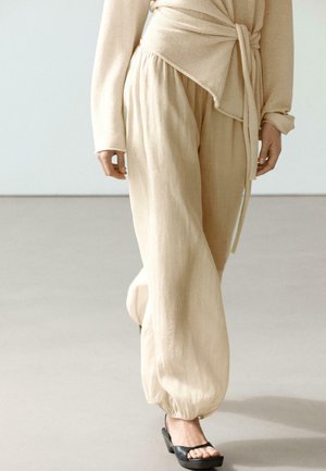 Beżowe luźne spodnie ze ściągaczami przy kostkach, w połączeniu z beżowym swetrem kopertowym i czarnymi sandałami na obcasie z odkrytymi palcami na neutralnej podłodze.