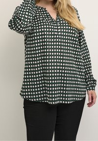 Groene en witte blouse met een patroon van ruiten, voorzien van een V-hals en lange mouwen, gemaakt van lichte stof. Zwarte broek.