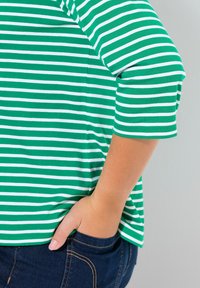 Grünes und weiß gestreiftes Oberteil mit Dreiviertelärmeln, weicher Stoff, lässige Passform, kombiniert mit dunkelblauen Jeans, Hand in der Tasche.