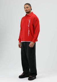 Rode poloshirt met lange mouwen met witte tekst "Young Holden", gecombineerd met wijde zwarte spijkerbroek en zwarte veterschoenen. Soepele stoftextuur.