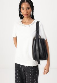 T-shirt blanc avec un col en perles, associé à un sac seau en cuir noir, doté d'une longue sangle. Un pantalon à pois noirs complète le look.