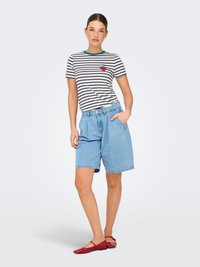 Mujer de pie con camiseta a rayas azul marino con parche de fresa, pantalones cortos de mezclilla azul claro y zapatos planos rojos sobre un fondo liso.