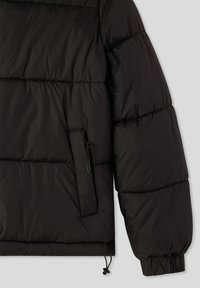 Maniche e lato di una giacca imbottita nera, con tasca con zip e polsino elastico, su uno sfondo grigio chiaro.