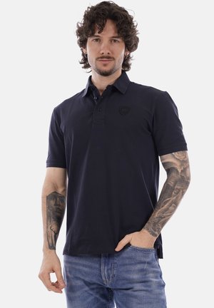 Uomo con capelli ricci che indossa una polo nera e jeans blu, mostrando tatuaggi dettagliati su entrambi gli avambracci, in piedi con una mano in tasca.