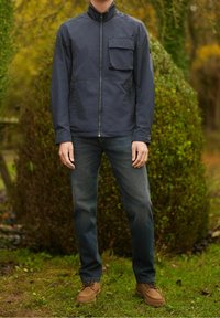 Marineblauw jack met rits en hoge kraag en een borstzak, gedragen over donkere jeans en lichtbruine schoenen, afgebeeld tegen een groene achtergrond.