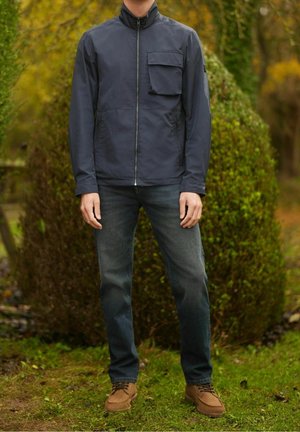 Marineblauw jack met rits en hoge kraag en een borstzak, gedragen over donkere jeans en lichtbruine schoenen, afgebeeld tegen een groene achtergrond.