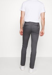 Šedé slim-fit kalhoty s hladkým povrchem, dvě zadní kapsy se zapínáním na knoflíky, v kombinaci s jednoduchým bílým tričkem a bílými teniskami.