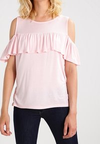 Jasnoróżowa bluzka z krótkim rękawem, z falbaną przy dekolcie i wycięciami na ramionach. Miękka tkanina o luźnym kroju i lekkim rozciągnięciu.