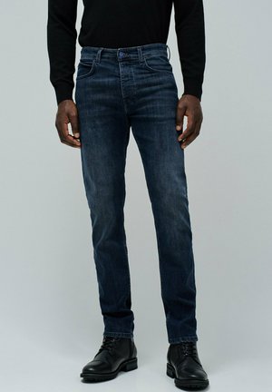 Jeans de mezclilla azul oscuro con un corte ajustado, que cuentan con un diseño estándar de cinco bolsillos y un cierre de cremallera.