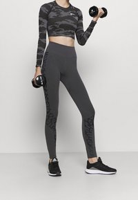 Grijze camouflage crop top en leggings met zwarte luipaardprint accenten. Model houdt zwarte dumbbells vast, draagt zwarte sportschoenen.