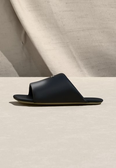 Μαύρο σανδάλι slide με ανοιχτά δάχτυλα, φαρδιά λωρίδα και επίπεδη σόλα, σε φόντο από μπεζ υφασμάτινη υφή.