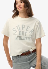 Cremefarvet t-shirt med rund hals, der har store trykte bogstaver "SUPER DRY ATHLETIC" i grå, korte ærmer og en afslappet pasform.