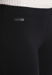 Detailní záběr na černé přiléhavé kalhoty s malou obdélníkovou etiketou značky Polo blízko pásku, které jsou nošeny s bílým žebrovaným topem.