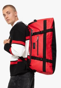 Sac à dos rouge et noir avec bretelles réglables, des zips, une poche avant et un design robuste. Texture lisse et bandes de couleur contrastantes.