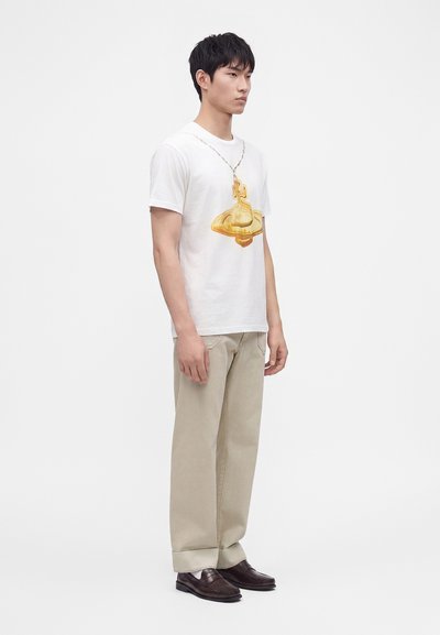 Νέος άνδρας στέκεται φορώντας λευκό t-shirt με στάμπα χρυσού φυλαχτού, μπεζ παντελόνι και σκούρα καφέ μοκασίνια σε απλό φωτεινό φόντο.