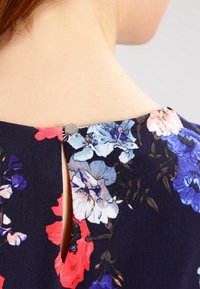 Blommönstrad tyg i nyanser av blått, rosa och rött, med en baksida som stängs med en metallknapp mot en mörk bakgrund.