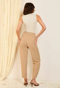 Beige high-waisted trousers made from a smooth fabric, featuring a tapered leg and side slits. Paired with strappy black sandals.