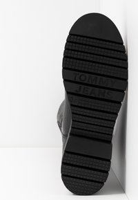 Semelle de botte en caoutchouc noir avec une adhérence texturée et à motifs. "TOMMY JEANS" embossé sur la zone du talon. Design plat et durable.
