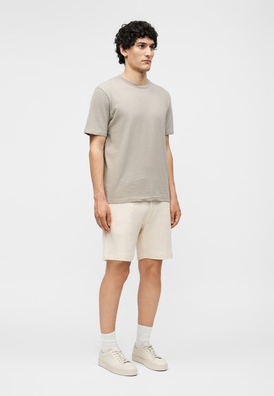 Άνδρας που στέκεται φορώντας μία μπεζ κοντομάνικη μπλούζα, κρεμ σορτς, λευκές κάλτσες και μπεζ αθλητικά παπούτσια μπροστά σε απλό ανοιχτό φόντο.