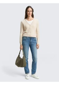 Cămașă cardigan bej cu zale, asociată cu blugi de culoare albastru deschis. Modelul poartă o geantă de umăr de culoare verde închis și adidași albi.