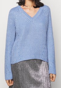 Modrý pletený svetr s výstřihem do V, se strukturou žebrování a dlouhými rukávy, doplněný stříbrnou plisovanou sukní.