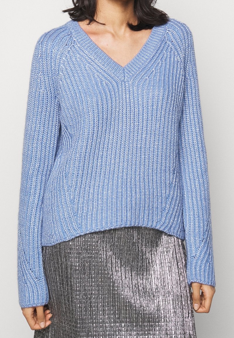 Modrý pletený svetr s výstřihem do V, se strukturou žebrování a dlouhými rukávy, doplněný stříbrnou plisovanou sukní.