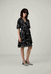 Rochie neagră cu motive circulare albe și accente distinctive, cu mâneci scurte, curea la talie și tiv cu detalii de marcă. Purtați cu pantofi gri.
