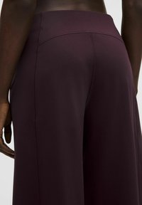 Detailný záber osoby v vysokom pásu, nosiacej tmavo fialové široké nohavice, zobrazuje detaily zadného a bočného švu.