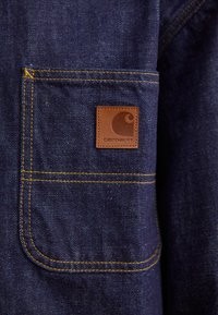 Chaqueta de mezclilla azul oscuro con costuras amarillas, que cuenta con un bolsillo en el pecho y un parche de cuero cuadrado en relieve con un diseño de ola.