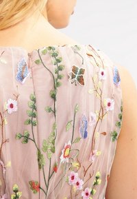 Ljust rosa genomskinlig tyg med intrikata blommönster i olika färger, med dragkedja bak och en texturerad design.