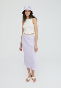 Witte cropped tanktop met zijkant ruches, lichtpaarse satijnen midi-rok, gestreepte paarse en witte bucket hat, en witte flip-flops.