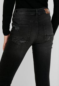 Jeans de mezclilla negros con una cintura alta, que presentan bolsillos traseros desgastados y una etiqueta de marca de cuero marrón. Textura suave, diseño ajustado.